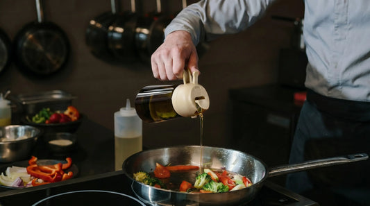 Person pouring oil from a kitchen oil sprayer into a pan with colorful vegetables including broccoli, bell peppers, and onions in a modern kitchen