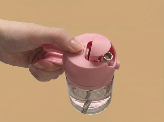 Hand holding a pink glass oil sprayer with the flip-top open against a clean beige background