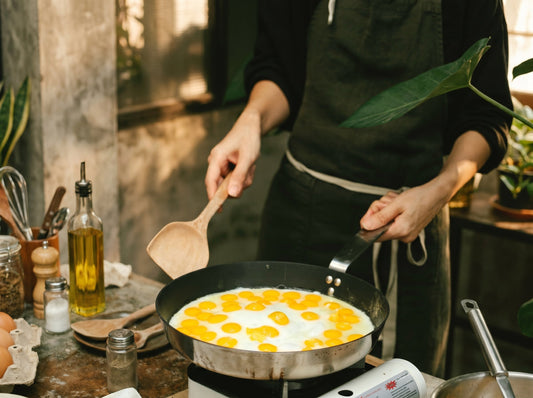 Person cooking eggs in a frying pan beside a glass olive oil dispenser in a cozy home kitchen