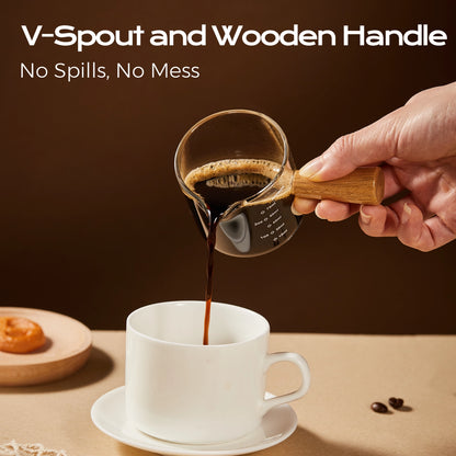 Espresso poured from the V-shaped spout of a wooden-handle glass measuring cup into a white mug.