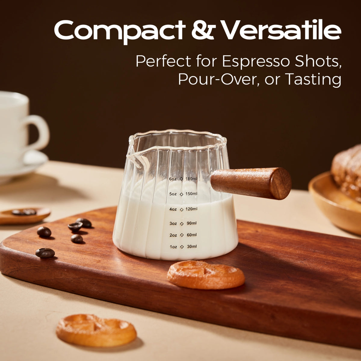 Fluted glass measuring pitcher filled with milk on a wooden board beside cookies and coffee beans.