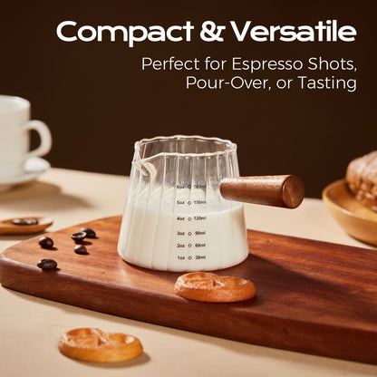 Fluted glass measuring pitcher filled with milk on a wooden board beside cookies and coffee beans.