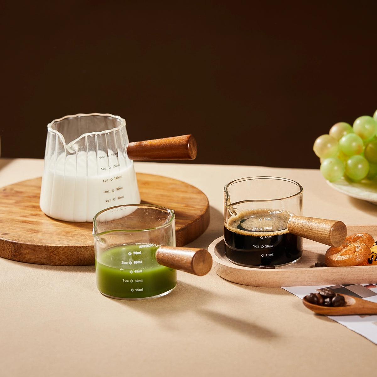 Wooden-handle glass pitchers holding milk, a green drink and espresso on a kitchen table.
