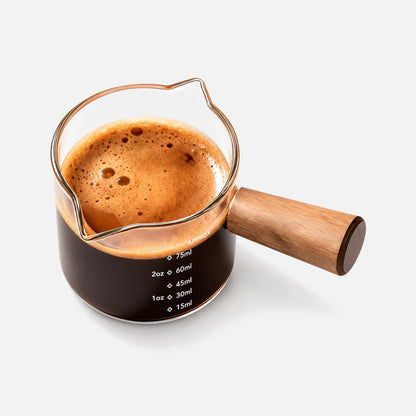 Glass espresso measuring cup with a wooden handle and dual pouring spouts.
