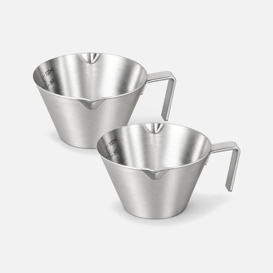 2 silver stainless steel espresso shot cups with handles and pouring spouts.